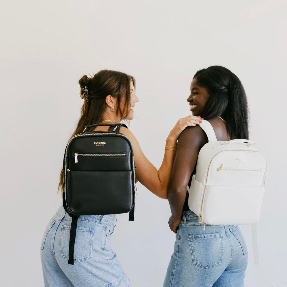 Two women smiling, wearing black and white backpacks.