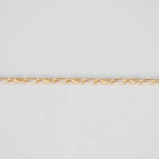 A close-up of a gold Figaro chain link against a plain light gray background.